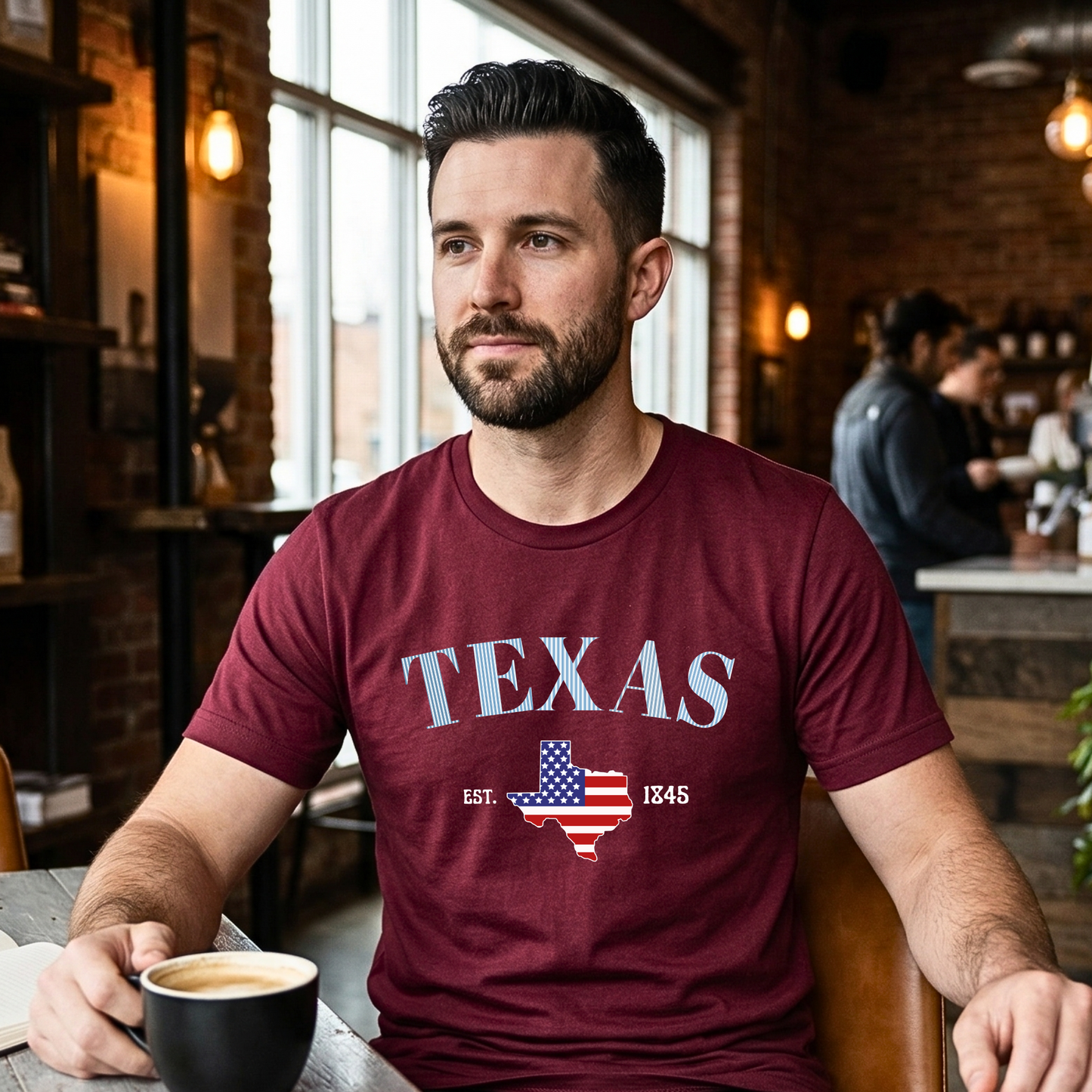 Man wearing a maroon 'TEXAS' t-shirt with an American flag design, sitting in a coffee shop.
