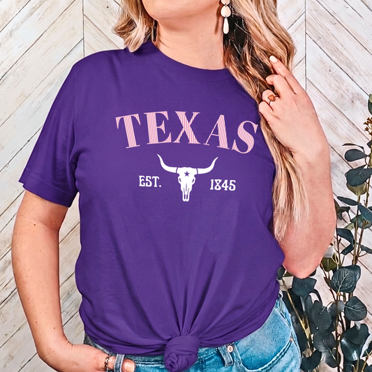 Person wearing a purple 'TEXAS' t-shirt with a skull design, standing against a wooden wall.