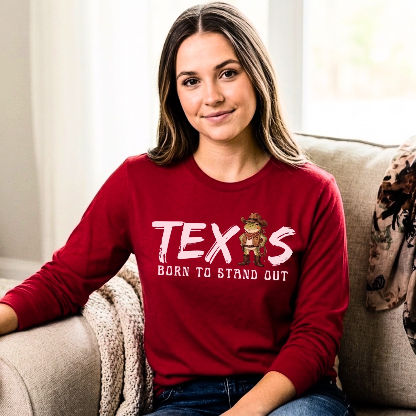Red long-sleeve t-shirt with the text 'TEXAS — Born to Stand Out' in white brush-style lettering, featuring a cartoon cowboy frog wearing a hat, bandana, and boots in place of the letter A. Shirt is shown on a smiling model seated on a couch in a bright, casual living room setting.