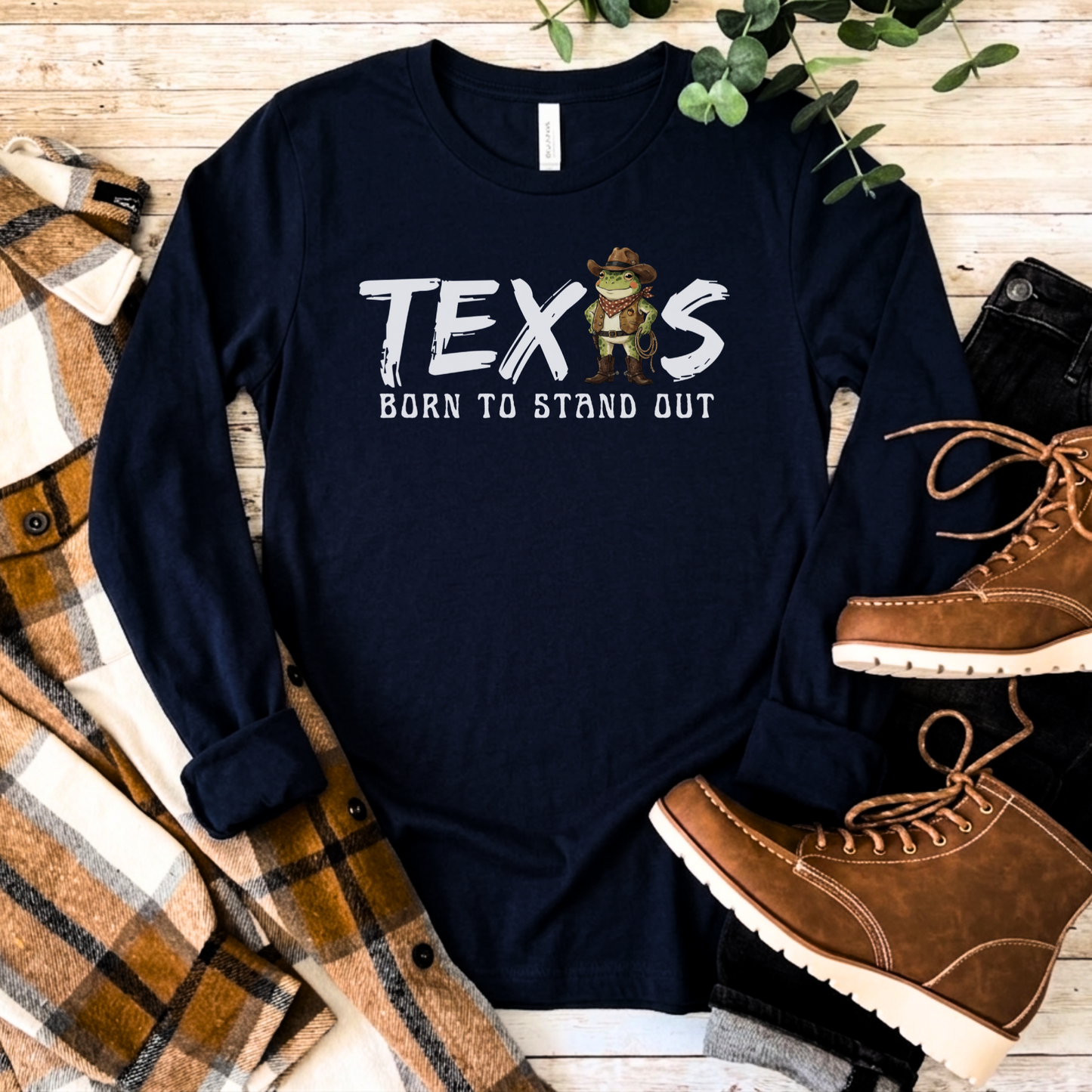 Navy blue long-sleeve t-shirt with the text 'TEXAS  Born to Stand Out' in white brush-style lettering, featuring a cartoon cowboy frog wearing a hat, bandana, and boots in place of the letter A. Shirt is displayed flat on a wooden surface surrounded by a plaid blanket and brown lace-up boots.
