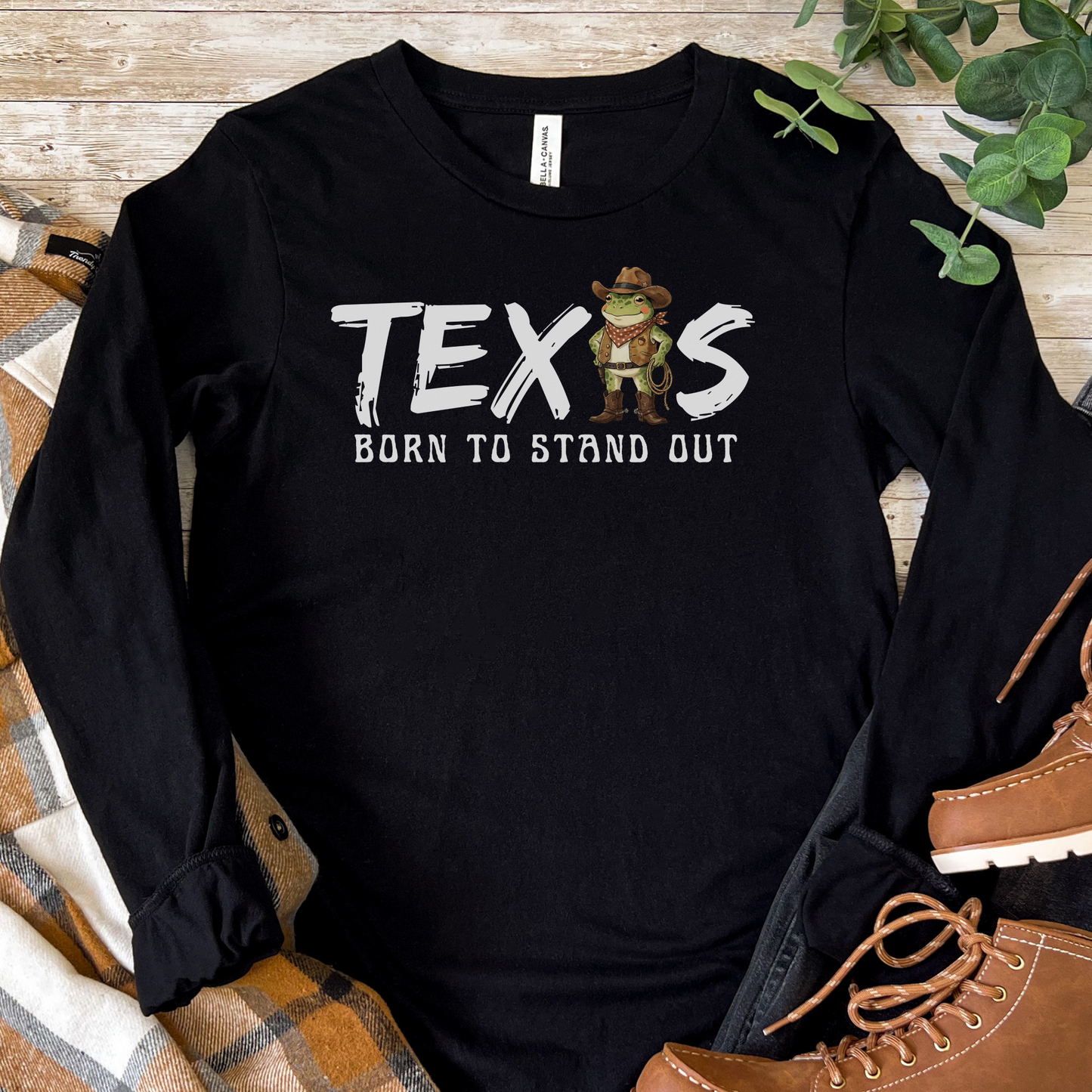 Black long-sleeve t-shirt with the text 'TEXAS — Born to Stand Out' in white brush-style lettering, featuring a cartoon cowboy frog wearing a hat, bandana, and boots in place of the letter A. Shirt is displayed flat on a wooden surface surrounded by a plaid blanket and brown lace-up boots.