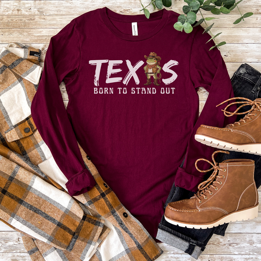 Maroon long-sleeve shirt with 'TEXAS Born to Stand Out' text, plaid blanket, and brown boots on a wooden surface.