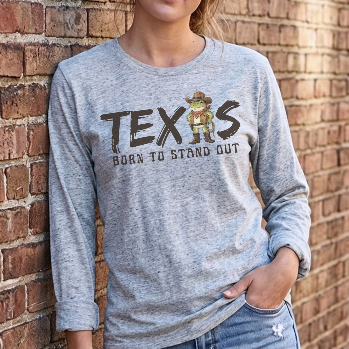 Gray long-sleeve t-shirt with the text 'TEXAS — Born to Stand Out' in dark brown brush-style lettering, featuring a cartoon cowboy frog wearing a hat, bandana, and boots in place of the letter A. Shirt is shown on a model leaning against a red brick wall