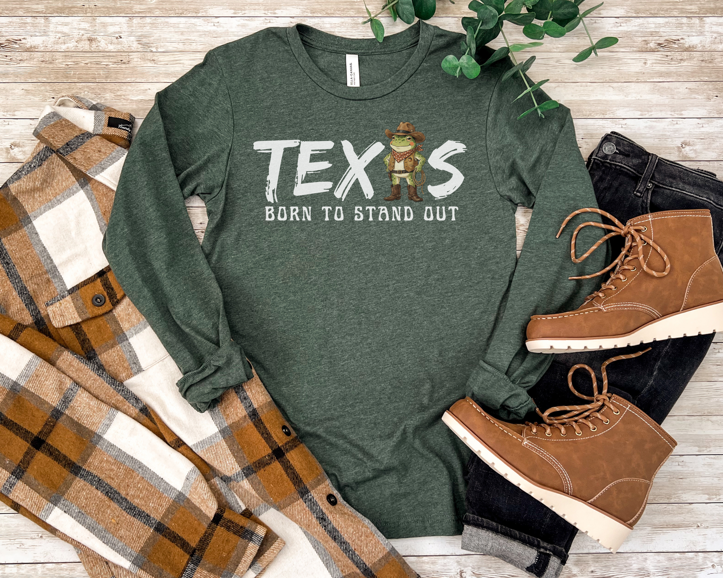 Green long-sleeve shirt with 'TEXAS BORN TO STAND OUT' text and graphics on a wooden surface with boots and plaid fabric.