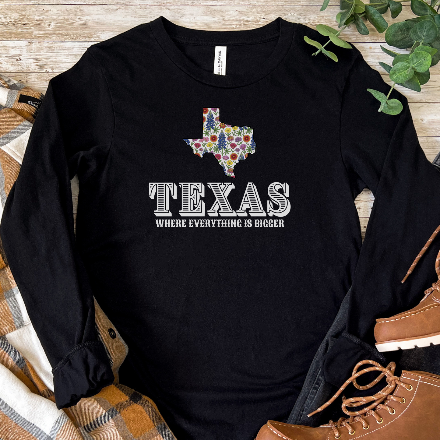 Black long-sleeve shirt with Texas design and text on a wooden surface with boots and plaid fabric.
