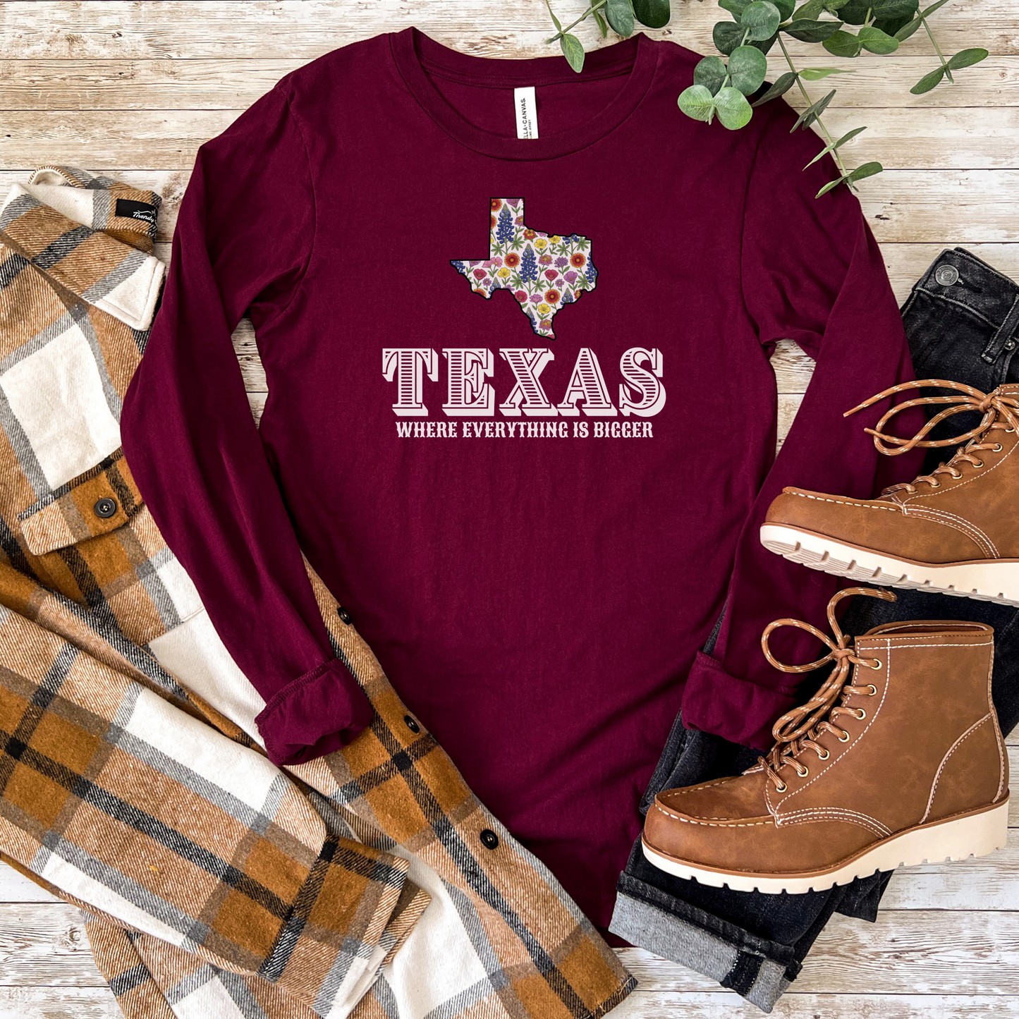 Maroon long-sleeve t-shirt featuring the outline of Texas filled with a colorful wildflower pattern above the text 'TEXAS  Where Everything Is Bigger' in white Western-style lettering. Shirt is displayed flat on a wooden surface surrounded by a plaid flannel and brown lace-up boots.