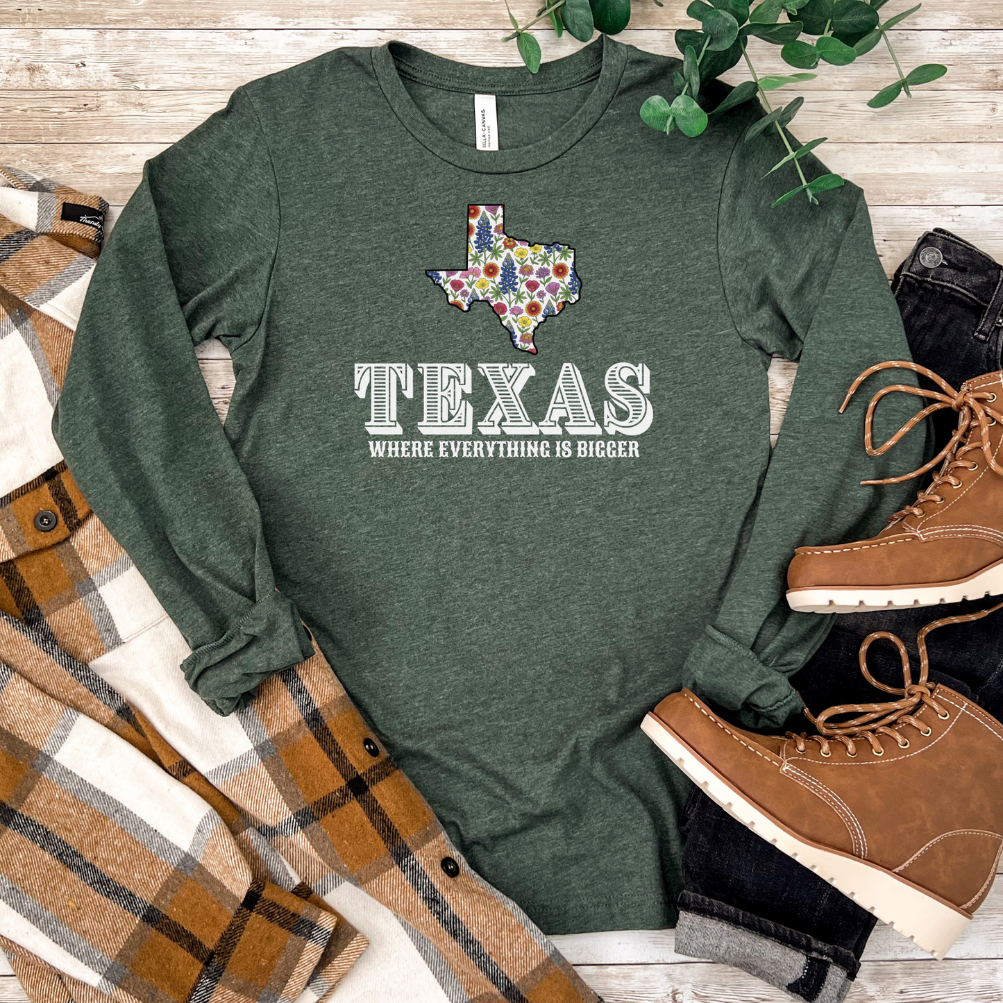 Green long-sleeve shirt with Texas design and text on a wooden surface with boots and plaid fabric.