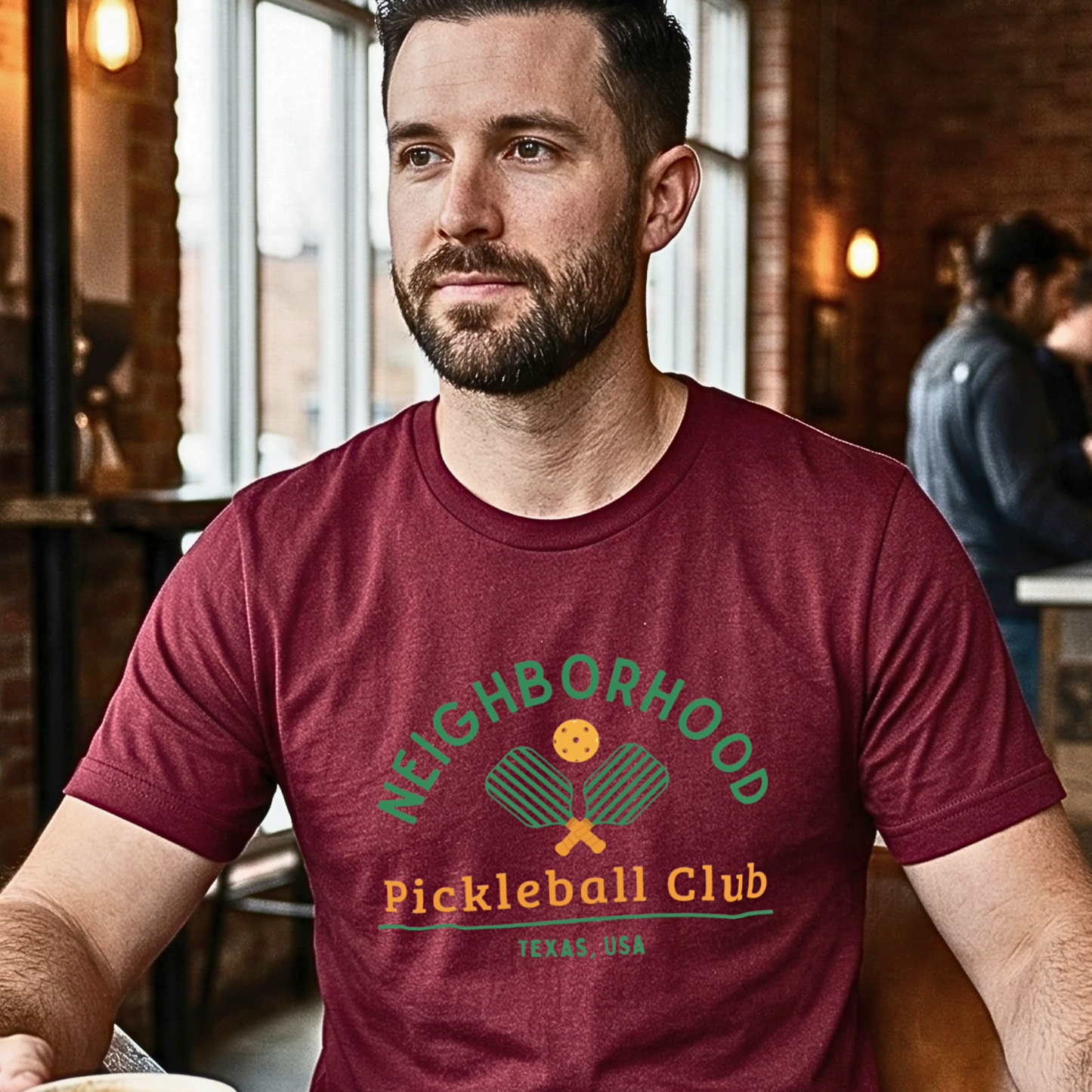 Man wearing a maroon t-shirt with 'Neighborhood Pickleball Club Texas, USA' design, sitting in a coffee shop.
