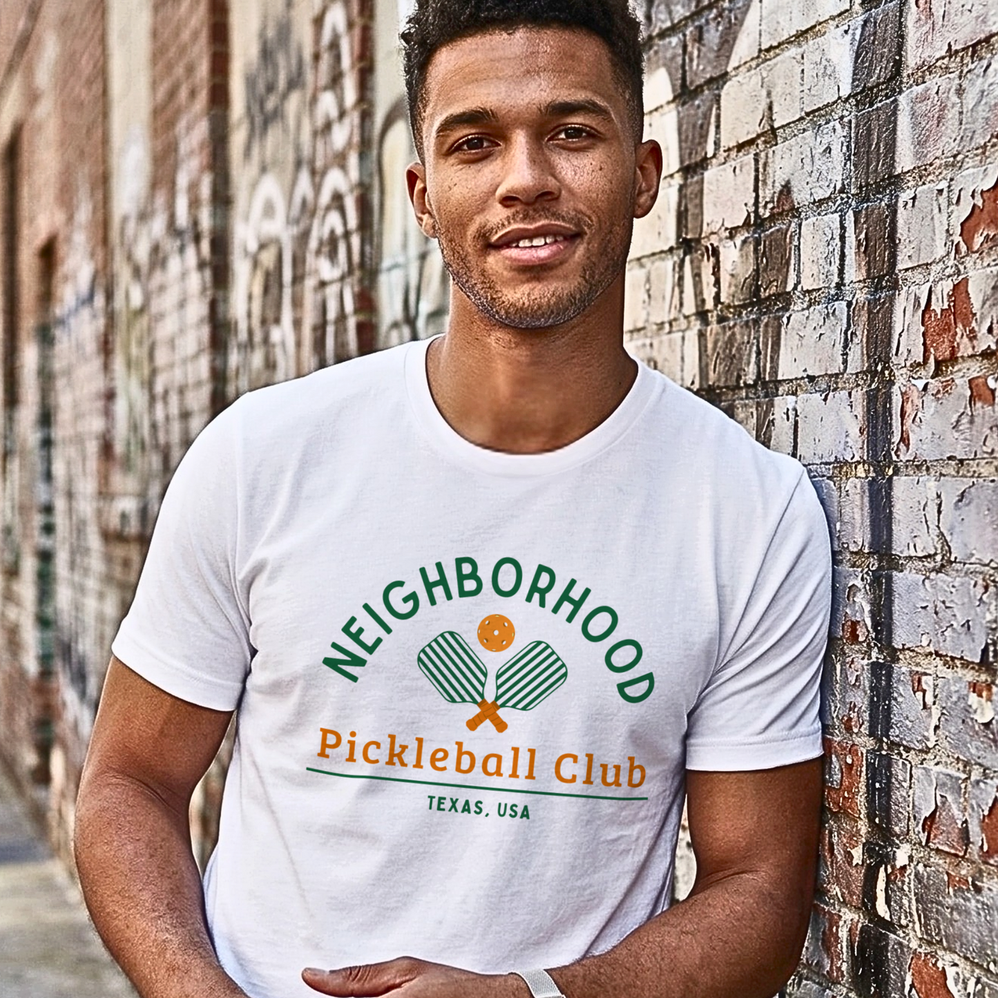 Man wearing a white t-shirt with 'Neighborhood Pickleball Club Texas, USA' design leaning against a brick wall.
