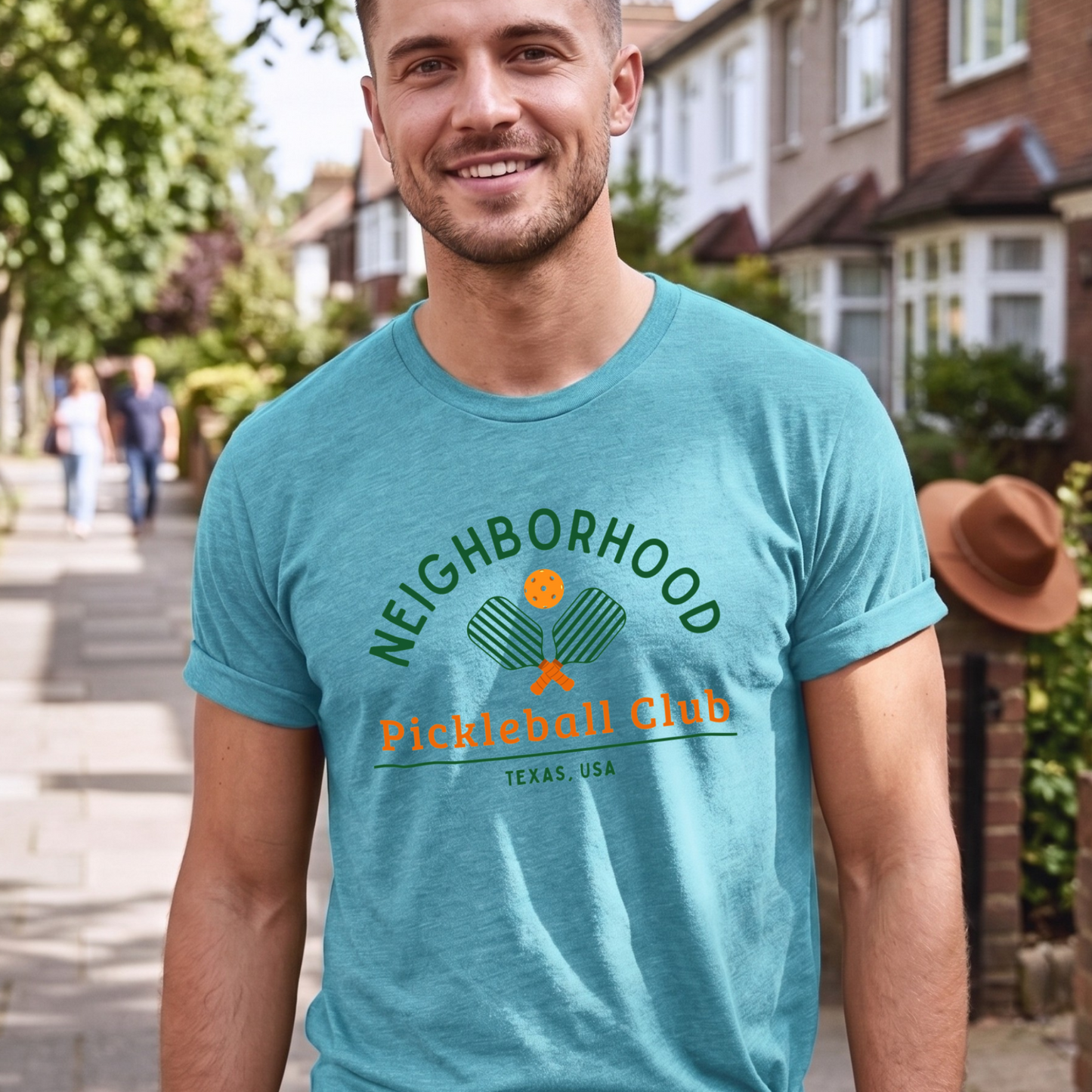 Man wearing a turquoise t-shirt with 'Neighborhood Pickleball Club Texas, USA' text and graphics, standing on a residential street.
